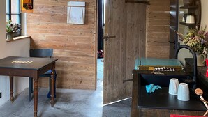 Een kamer met een houten wand, een eettafel, een stoel, een hanglamp, een deur en een wastafelgedeelte met bekers en een rode container.