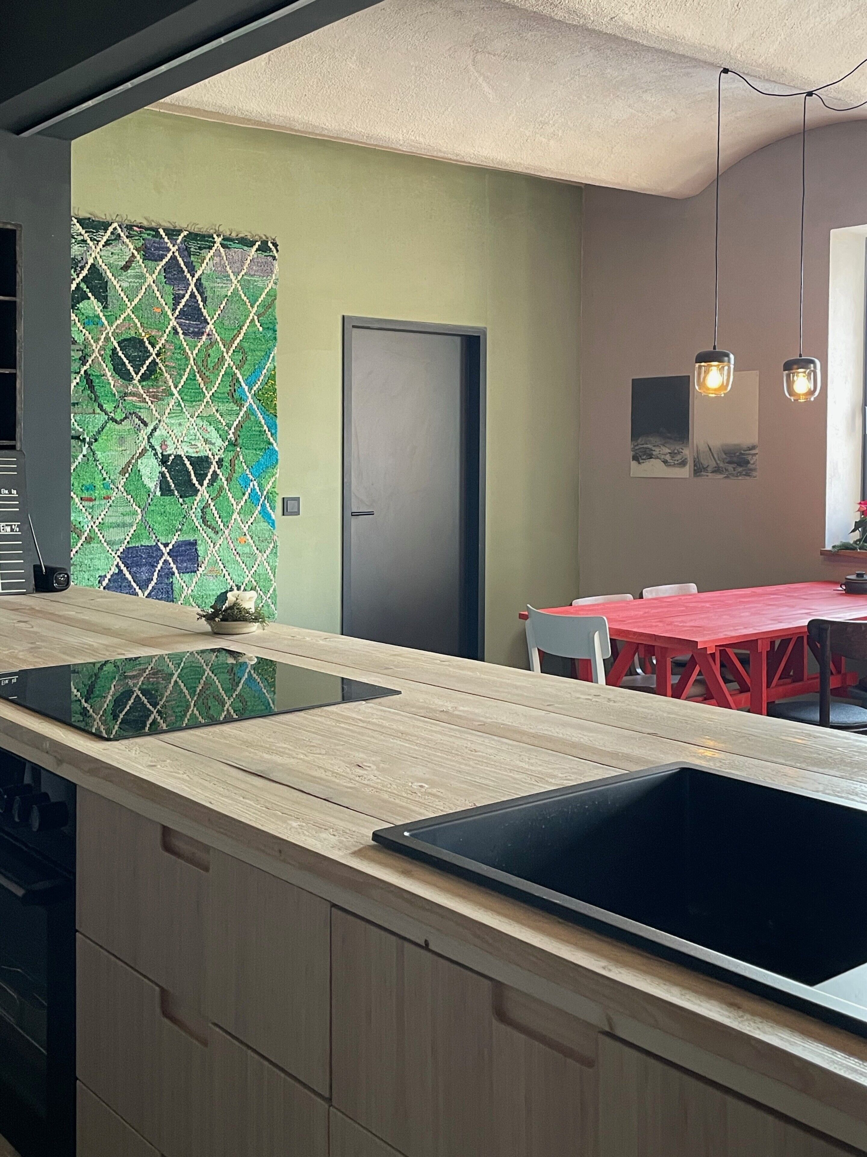 A modern kitchen with a large island, a dining area with red chairs, and a door with a green mosaic wall.