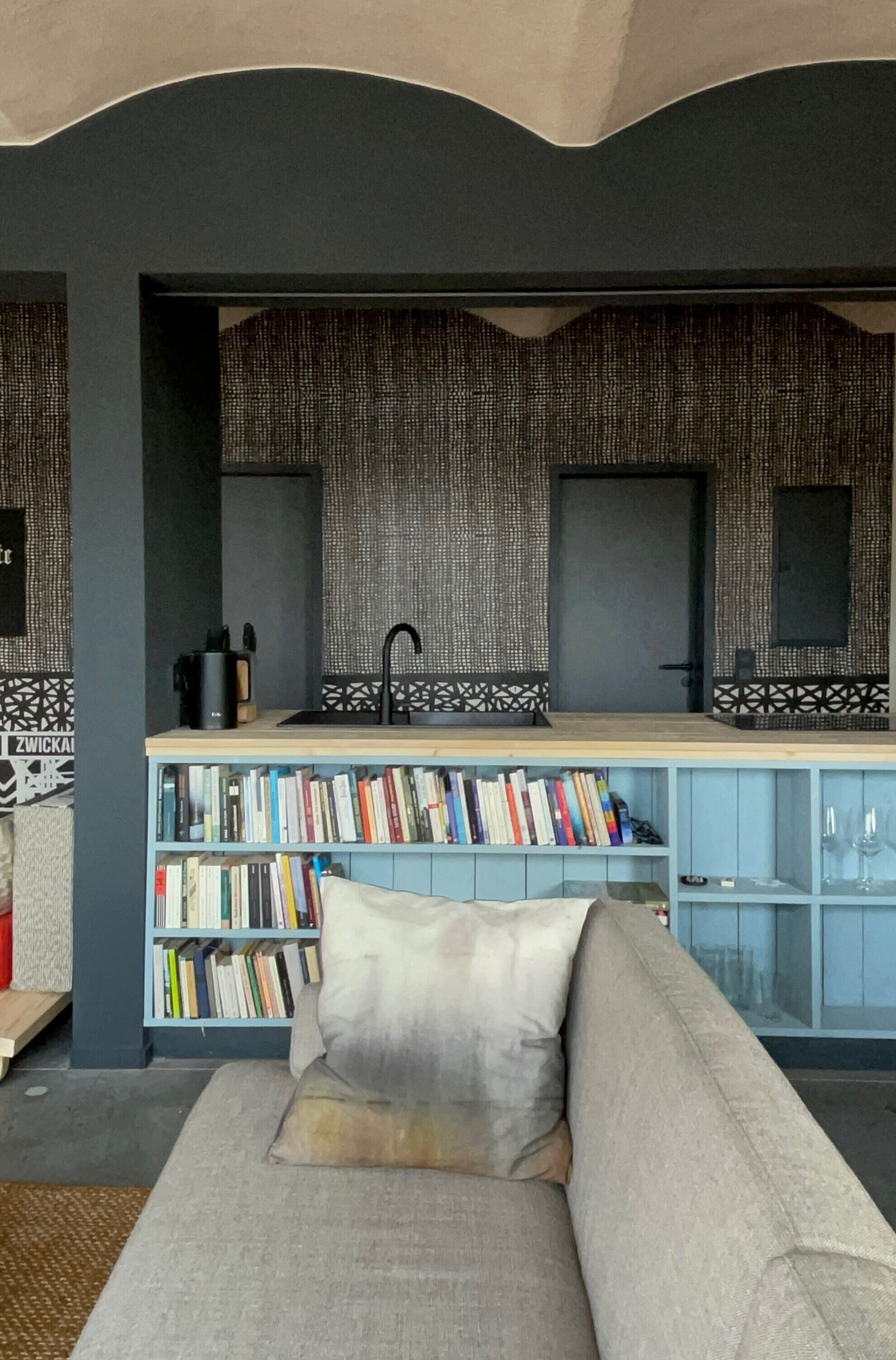 Un salon moderne avec un canapé, une bibliothèque remplie de livres, un coin cuisine avec un évier et un mur orné d’un motif.