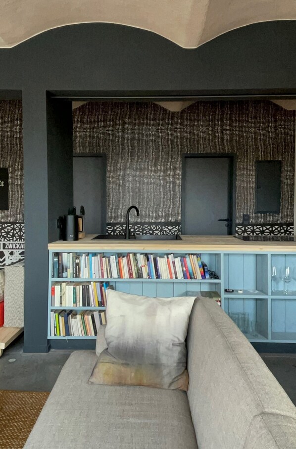 A modern living room with a sofa, a bookshelf filled with books, a kitchen area with a sink, and a patterned wall.