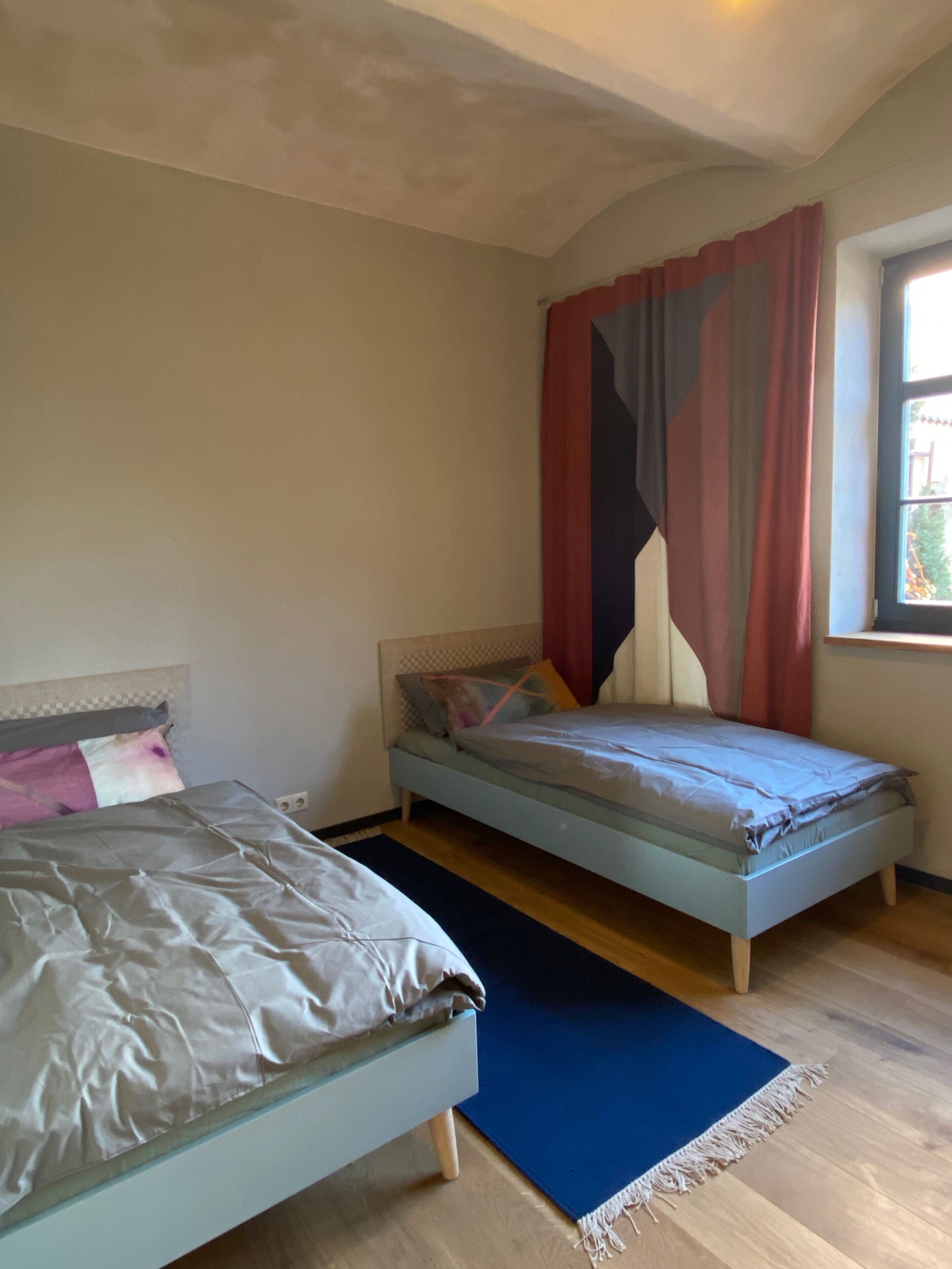 Een slaapkamer met twee bedden, een raam met gordijnen en een blauw vloerkleed.
