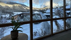 Ein Zimmer mit großem Fenster mit Blick auf eine verschneite Landschaft, Berge und eine Holzhütte.