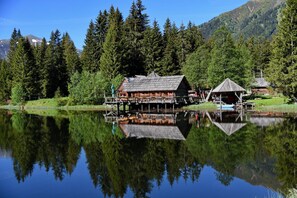 A wooden cabin with a dock extending into a calm lake, surrounded by dense forest and mountains.