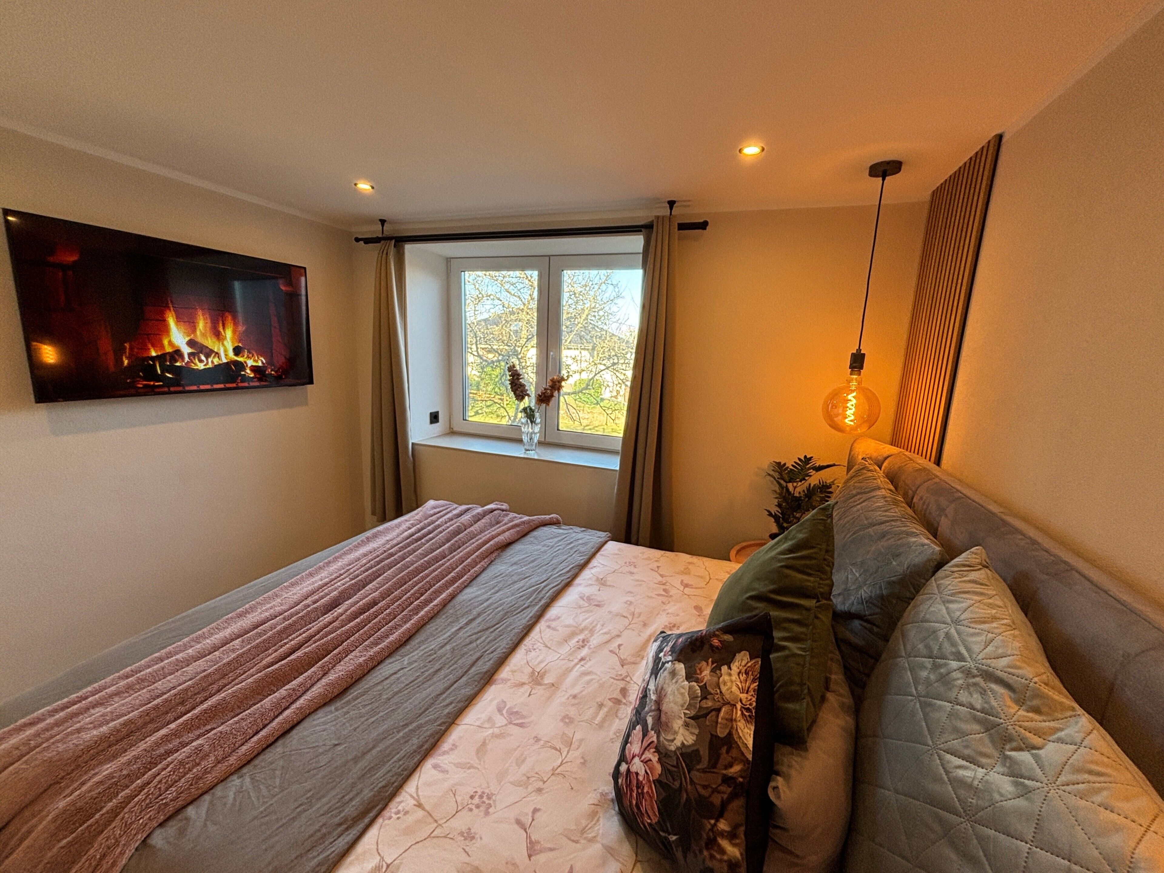 Une chambre à coucher avec un lit, une cheminée, une fenêtre avec des rideaux et un luminaire suspendu.