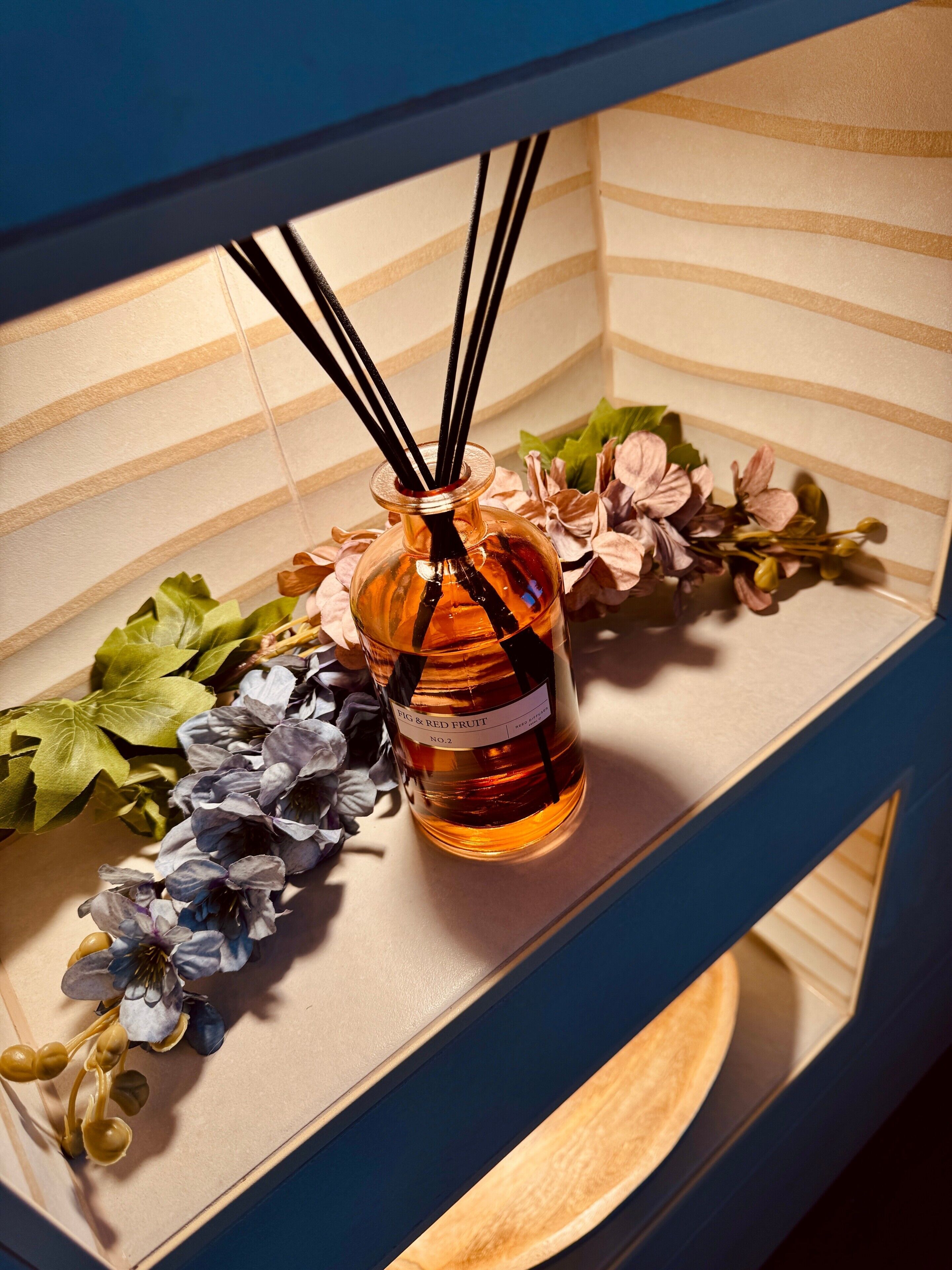 Een fles Fig & Red Fruit No.2 op een plank met decoratieve bloemen en een gestreepte achtergrond.