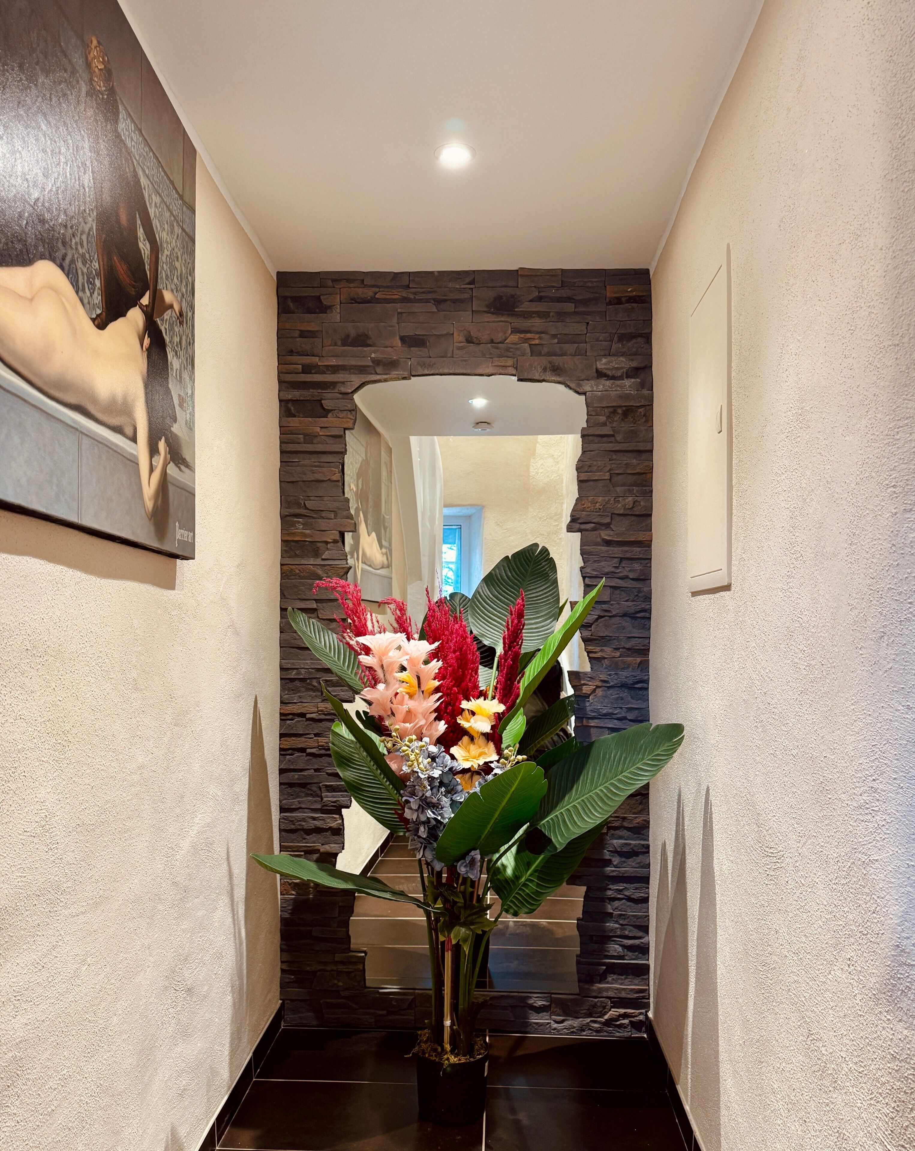 Een gang met een stenen muur, een lijst met kunstwerk, een potplant met bloemen en een spiegel die een kamer weerspiegelt.