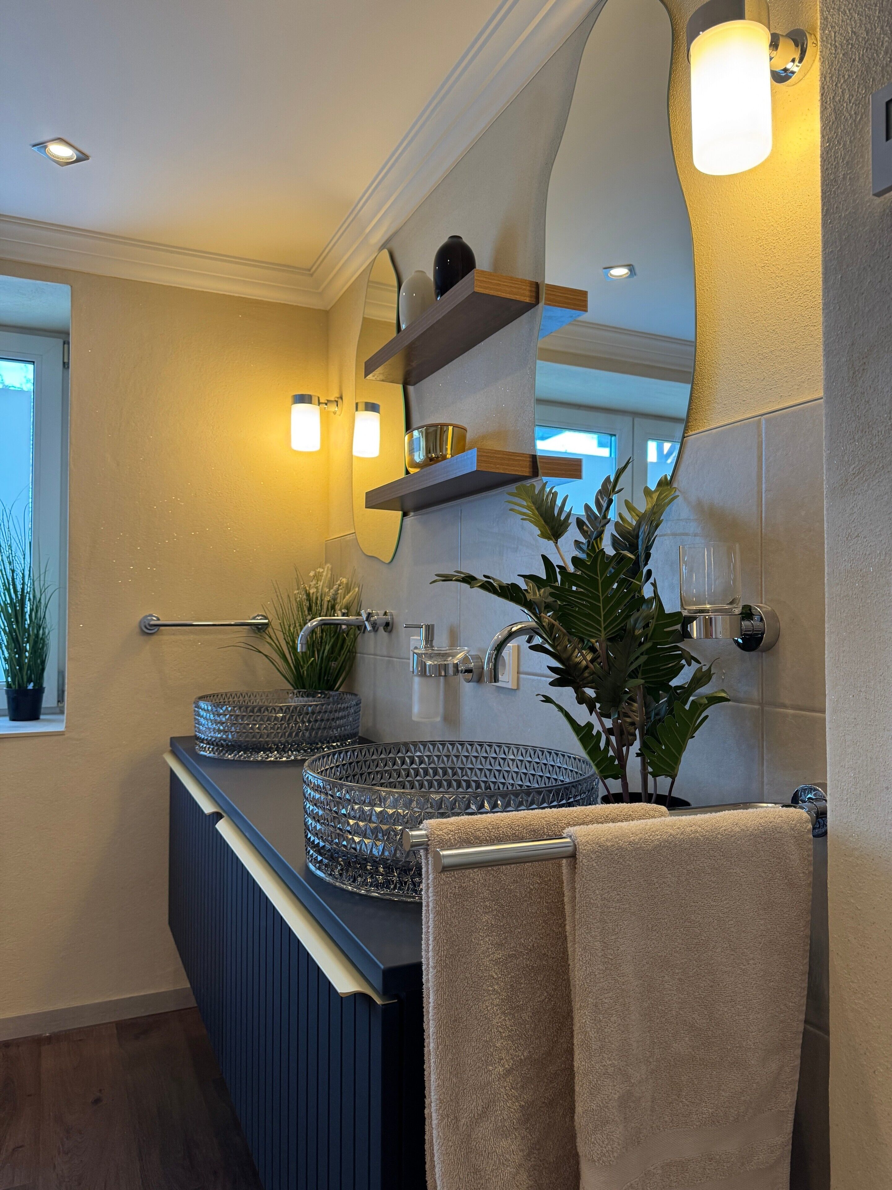 Une salle de bain moderne avec un lavabo en verre, un miroir avec des étagères, un porte-serviettes et une plante en pot.