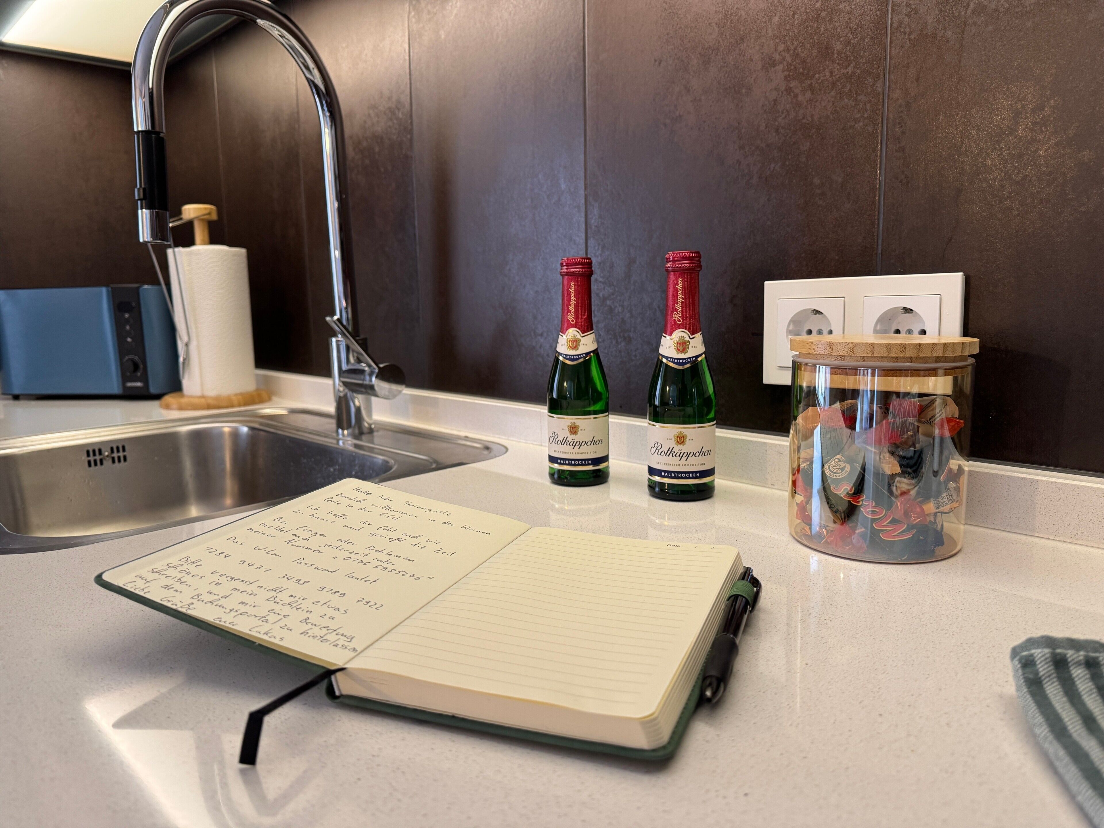 A kitchen counter with a notebook, two bottles of wine, a jar with items, and a faucet.