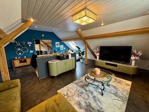 Uma sala de estar estilo loft com teto alto, vigas de madeira aparentes, uma TV de tela plana grande e uma área de jantar com mesa e cadeiras de madeira.