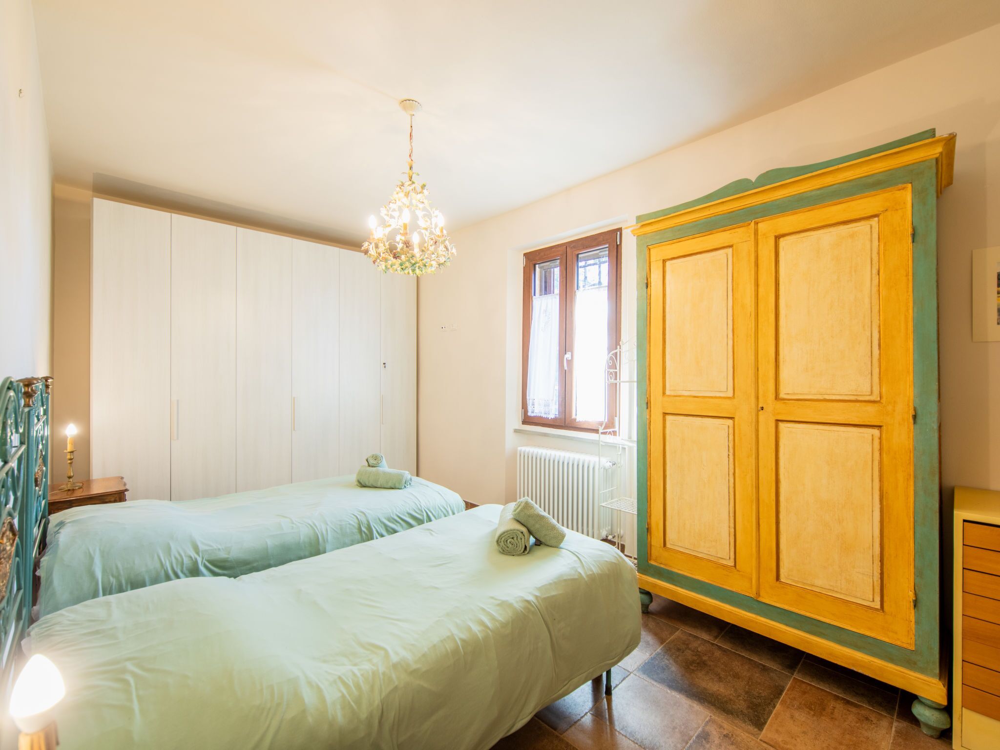 A bedroom with two beds, a chandelier, a wardrobe, and a window with curtains.