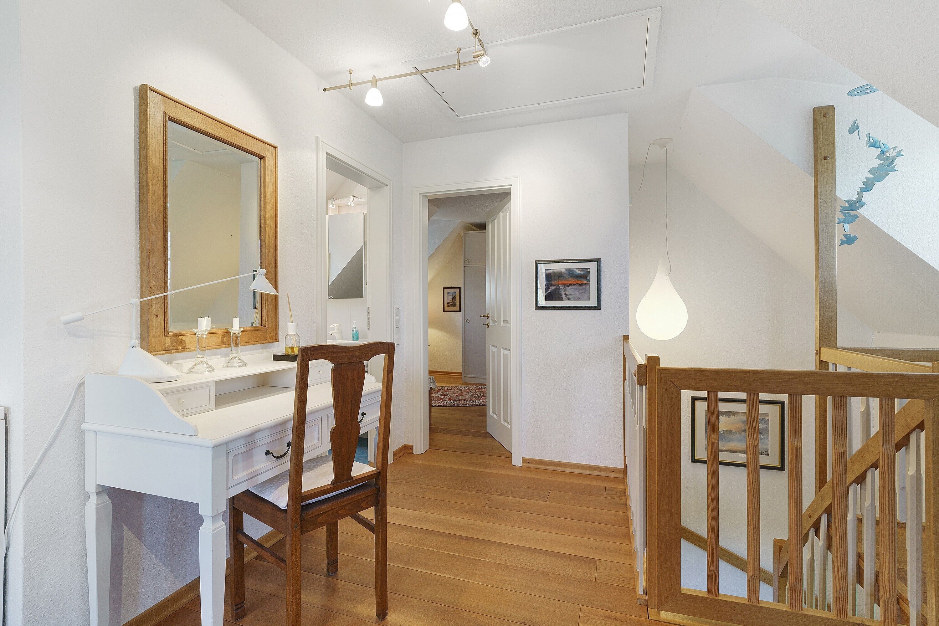 Un couloir doté d’un plancher en bois, d’un bureau blanc avec un miroir, d’une chaise en bois et d’un escalier avec une rampe en bois.