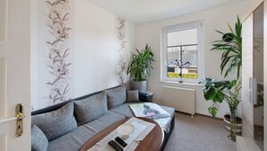 Una sala de estar con un sofá gris, una mesa de centro con un mantel individual, una planta en maceta y una ventana con vista a un balcón.