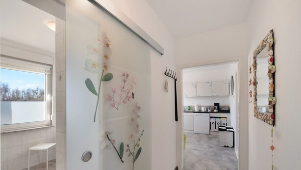 Un pasillo con un banco blanco, una puerta de cristal decorada con motivos florales, un perchero y una zona de cocina con armarios blancos y un lavabo.