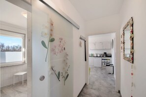 Un couloir avec un banc blanc, une porte en verre ornée de motifs floraux, un porte-manteau et un coin cuisine avec des placards blancs et un évier.