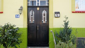 Una puerta doble negra con una pequeña ventana encima, flanqueada por dos números de casa, 14 y 12, sobre un muro amarillo y verde.