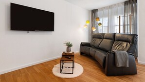 Een moderne woonkamer met een flatscreen-tv, een leren bank, een klein houten bijzettafeltje met een plant en een vloerlamp.