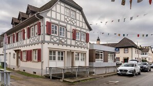 Een traditioneel vakwerkhuis met rode luiken, een hek en geparkeerde auto's langs de straat.