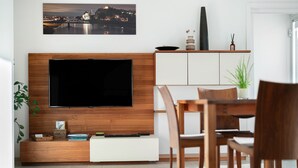 A modern living room with a flat-screen TV, wooden furniture, a dining area, and a cityscape painting.