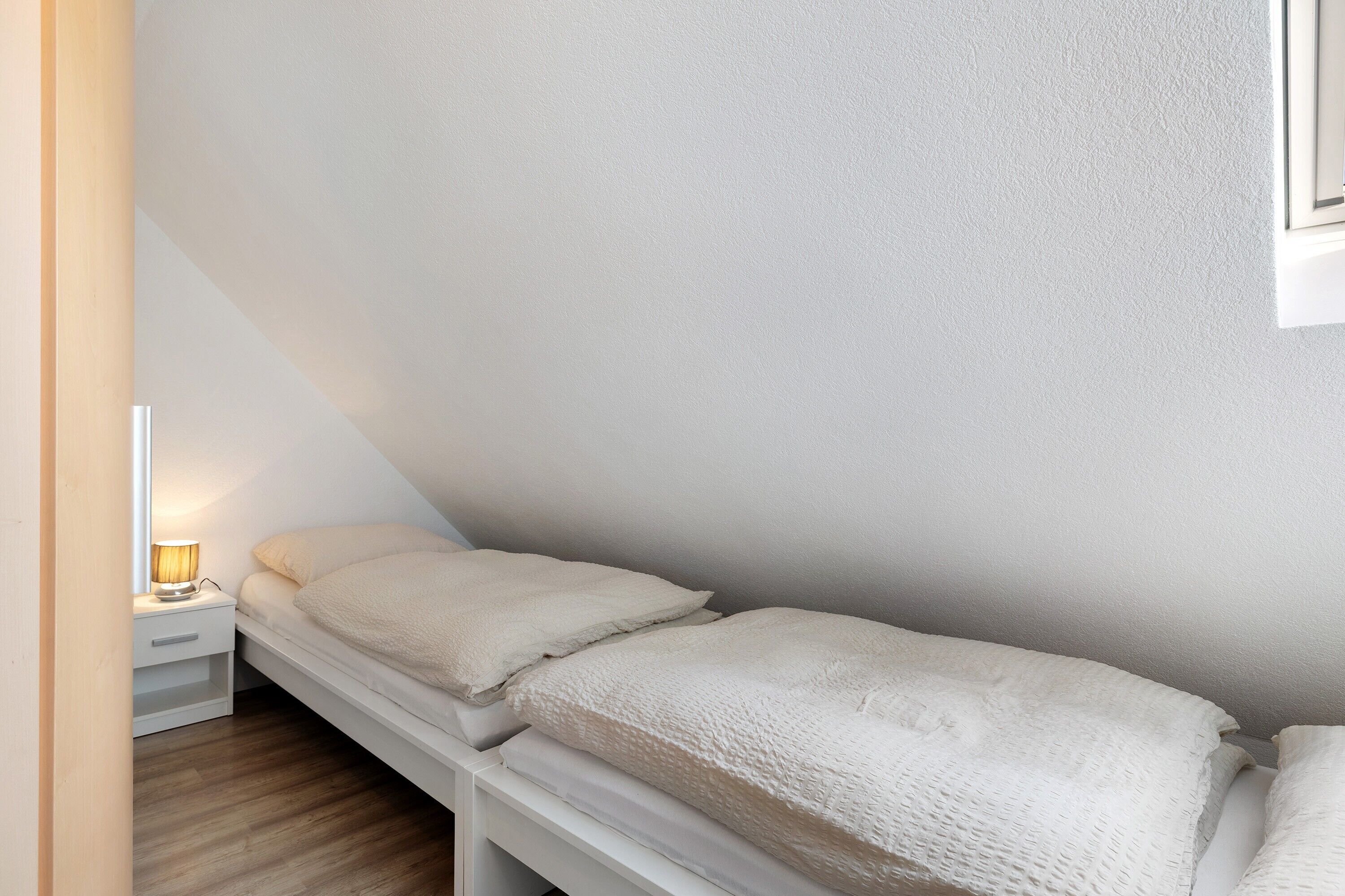 A small, single-bed room with a white bed, a bedside table with a lamp, and a white wall.