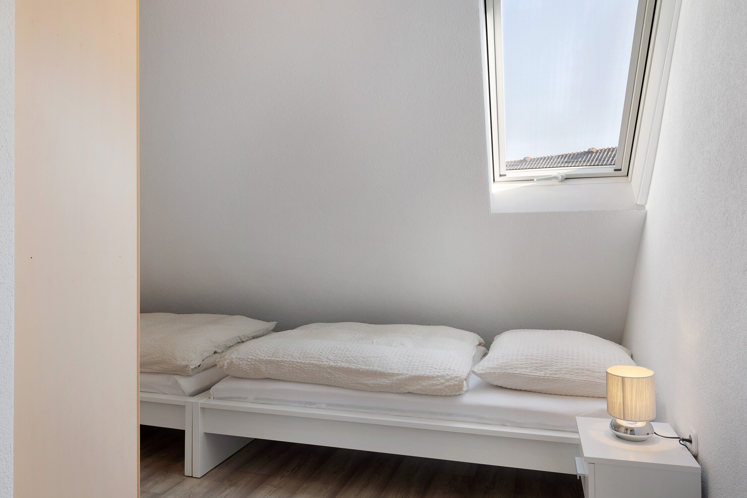 A small, white, single-bed room with a skylight and a bedside lamp.