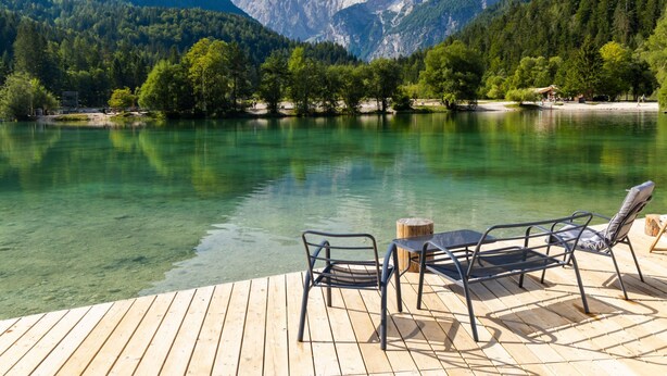 Een houten terras met metalen stoelen en een tafel, met uitzicht op een helder meer en bergen op de achtergrond.