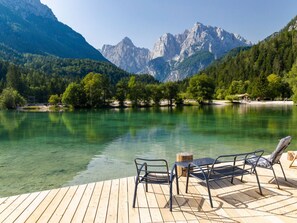 Eine Holzterrasse mit Metallstühlen und einem Tisch mit Blick auf einen klaren See, im Hintergrund Berge.