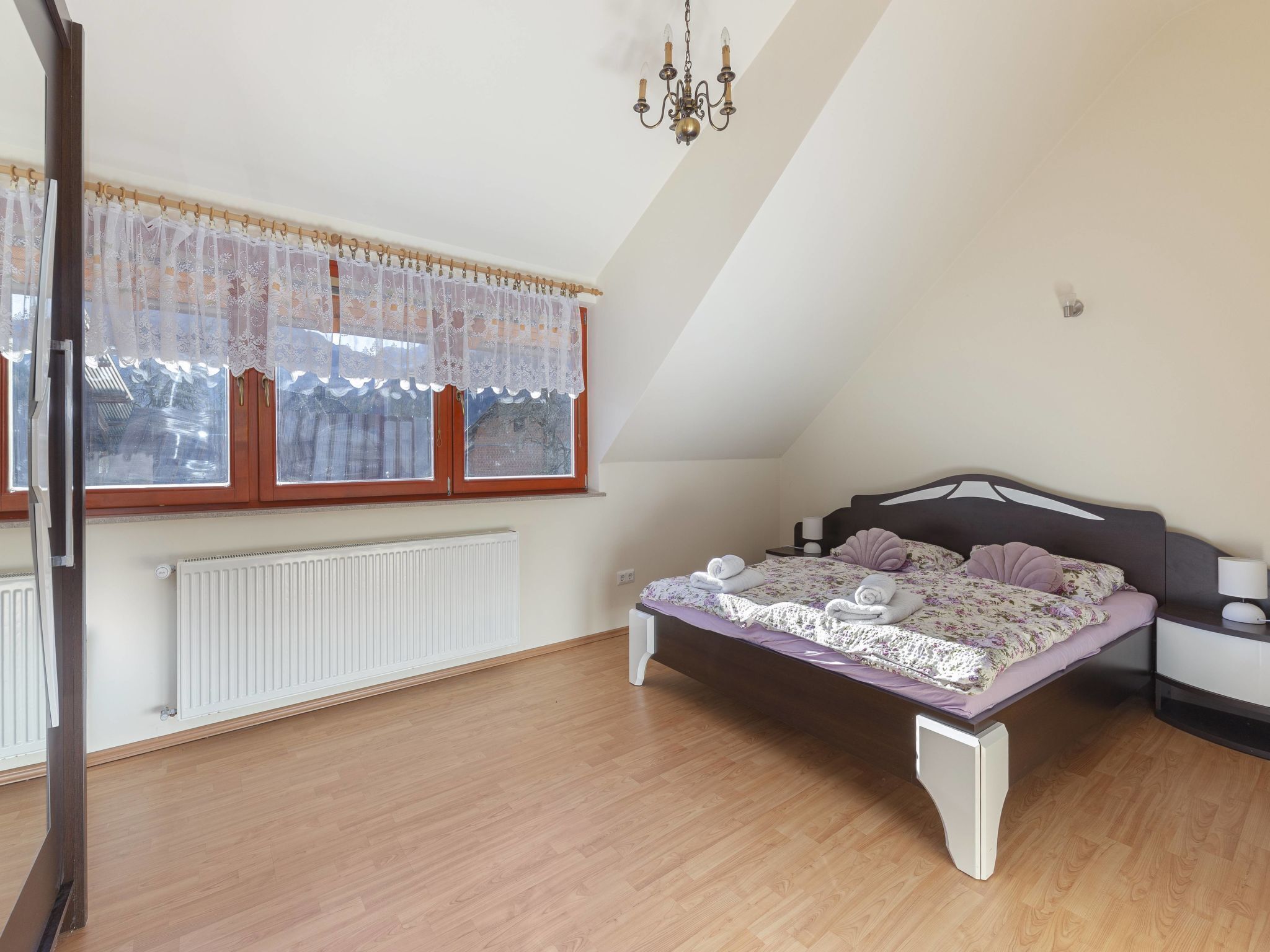 A bedroom with a sloped ceiling, a bed with a headboard, a window with lace curtains, a radiator, and a chandelier.