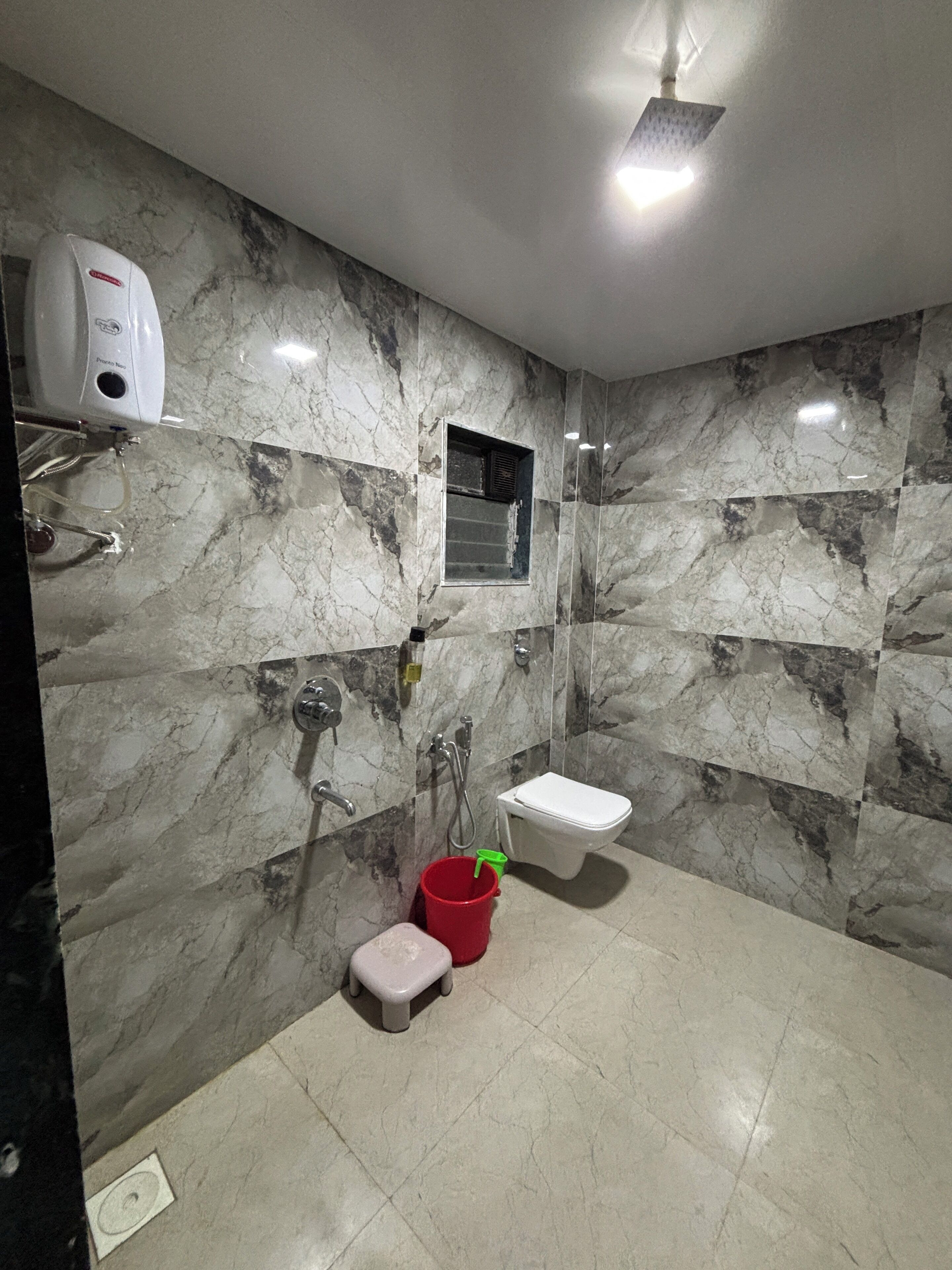 A bathroom with a marble wall, a white toilet, a red bucket, and a handheld showerhead.