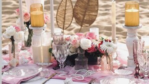 Una mesa frente al mar decorada con flores rosa y blancas, velas y cristalería.