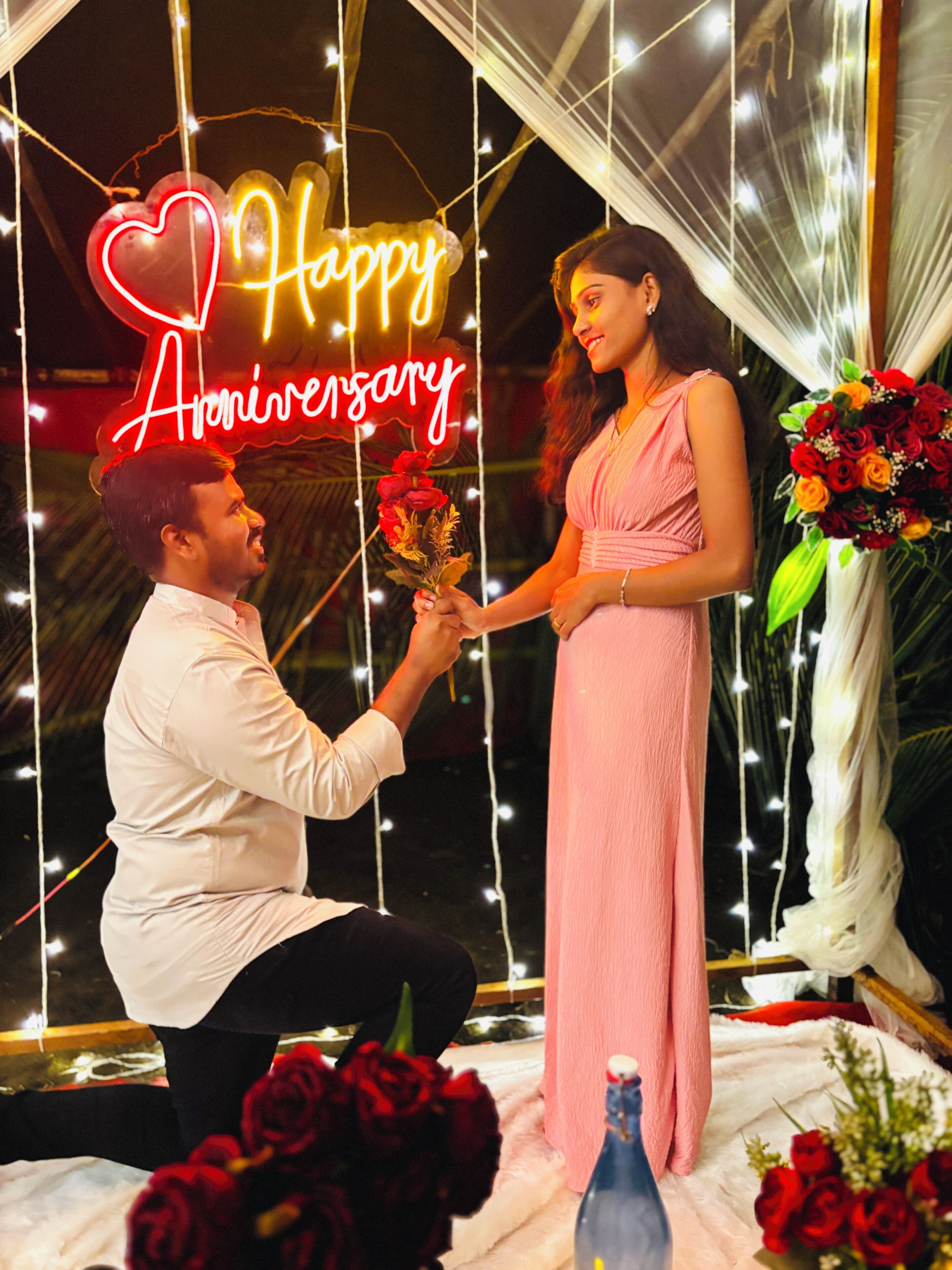 A man kneeling to propose to a woman with a 'Happy Anniversary' sign in the background.