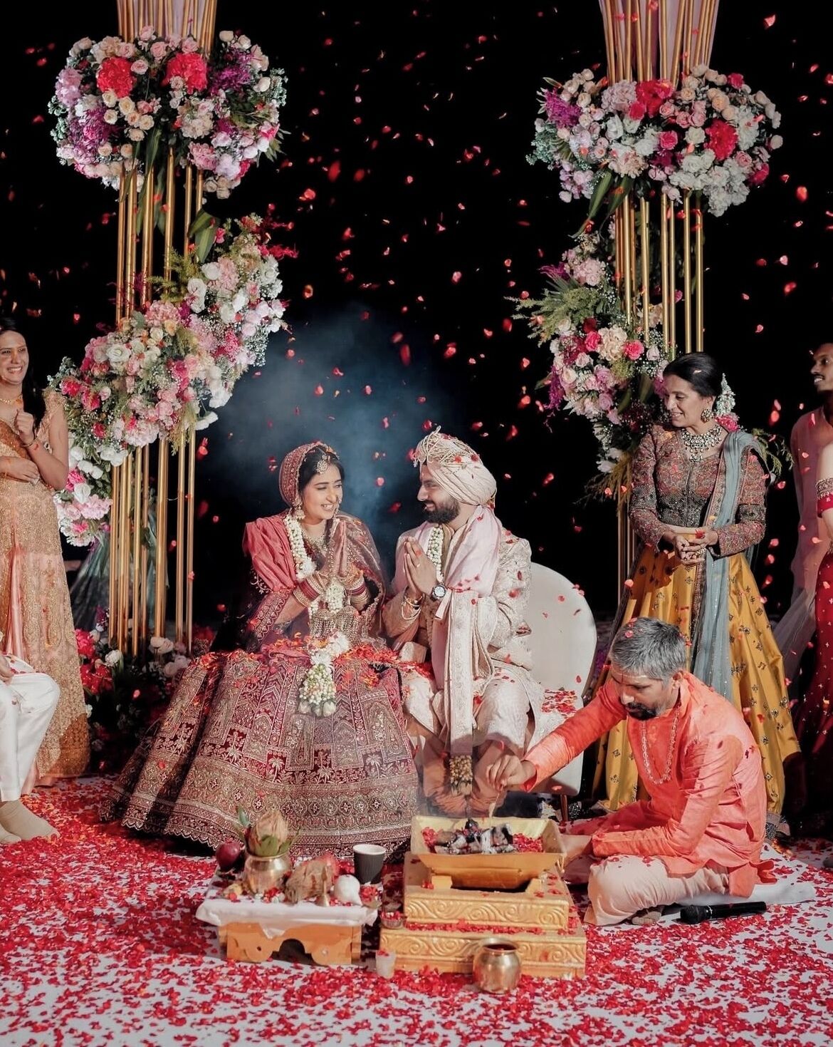 A traditional ceremony with participants seated and standing around a decorated area with floral arrangements and a central ritual setup.