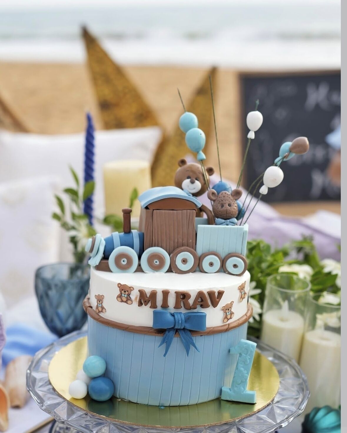 Un pastel de varios niveles con un diseño de oso y el nombre 'MIRAV' grabado, sobre un soporte de vidrio para pasteles.