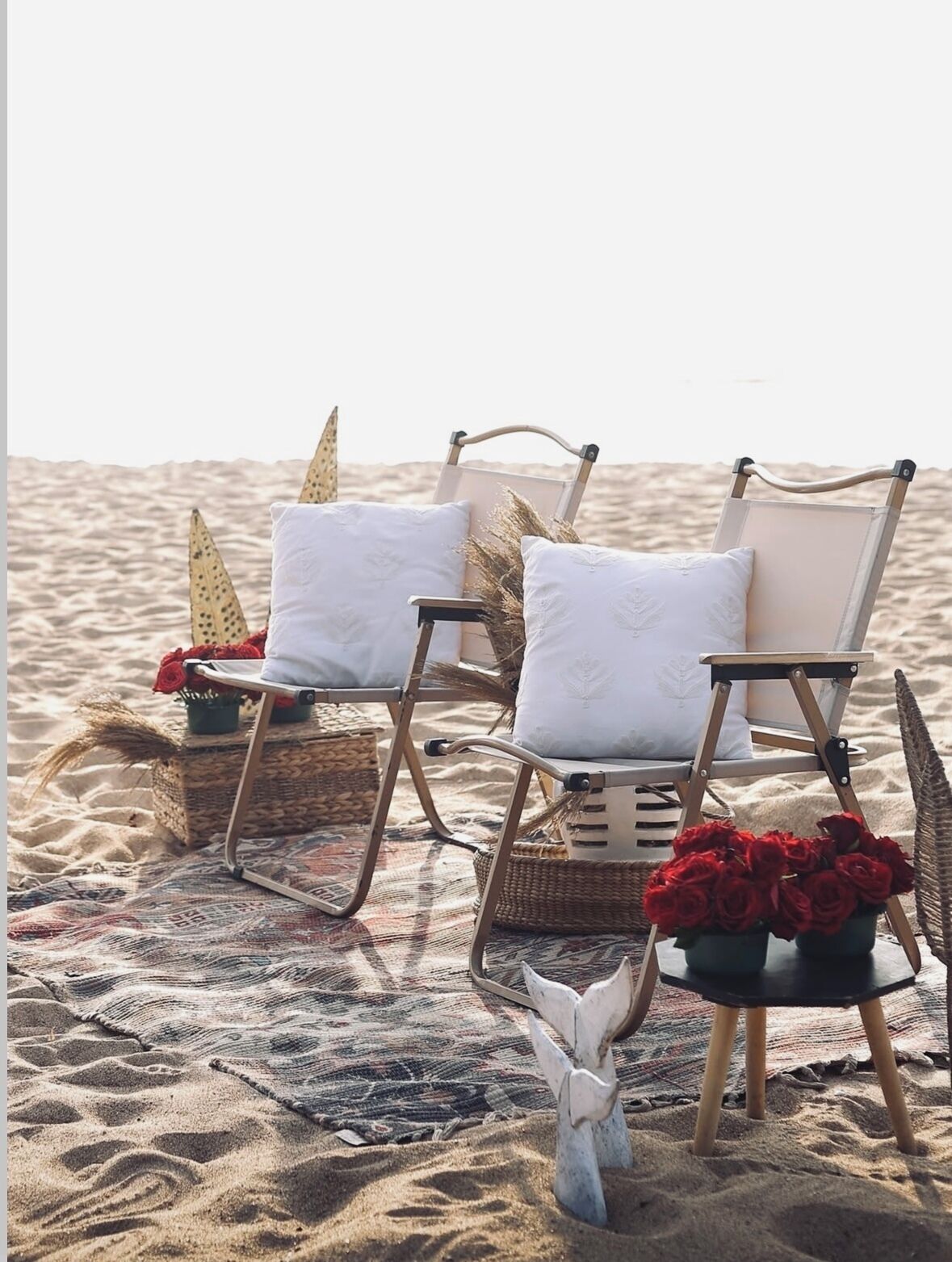 Two beach chairs with white cushions and decorative elements on a patterned rug, placed on a sandy beach.