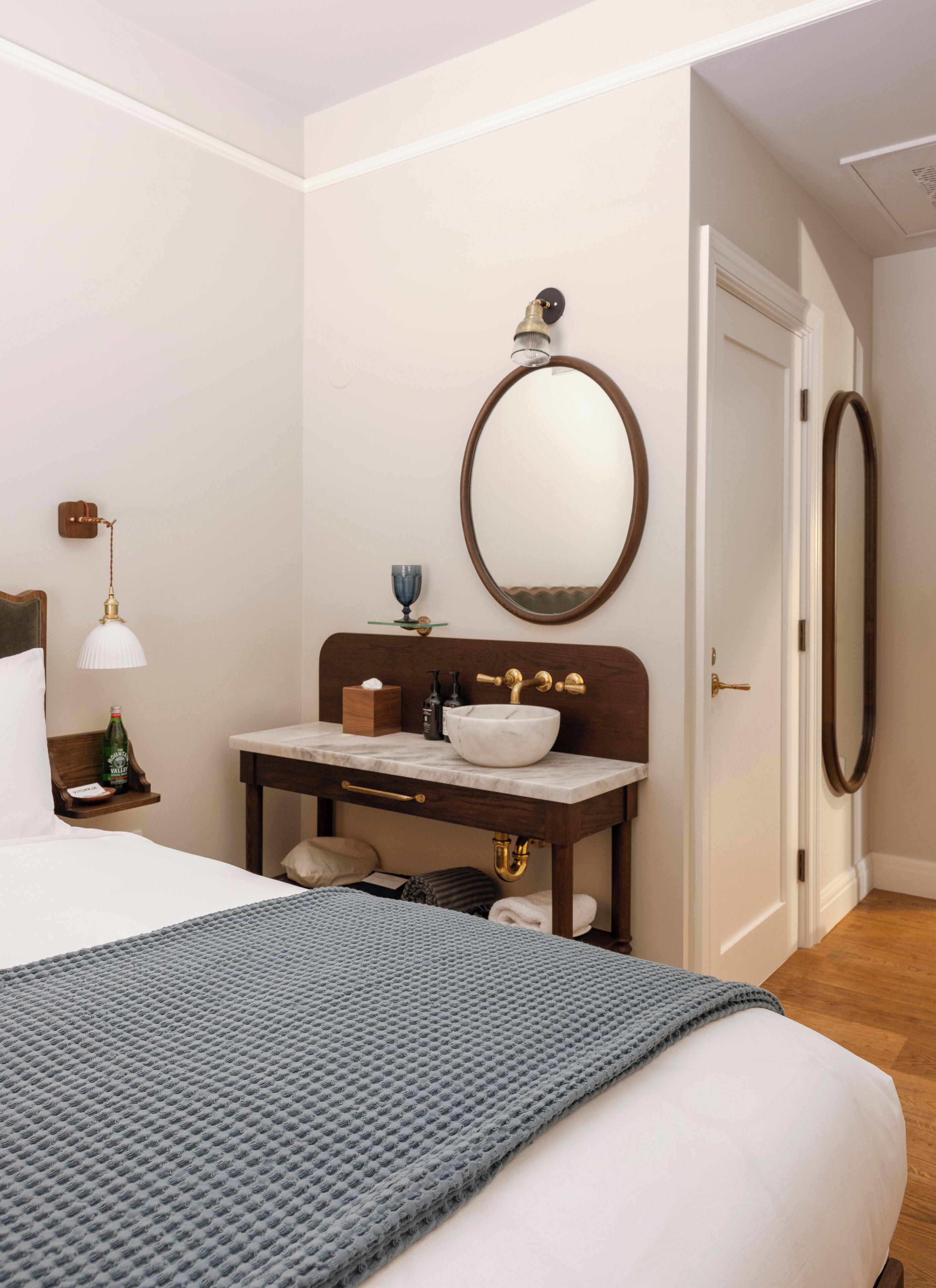 Une chambre d’hôtel comprenant un lit, une table de chevet avec une lampe, un miroir, une salle de bain avec un lavabo et une porte.