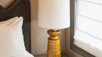 Un lit bien fait, avec du linge de lit blanc, une table de chevet en bois sur laquelle se trouve une lampe, et une fenêtre avec des stores.