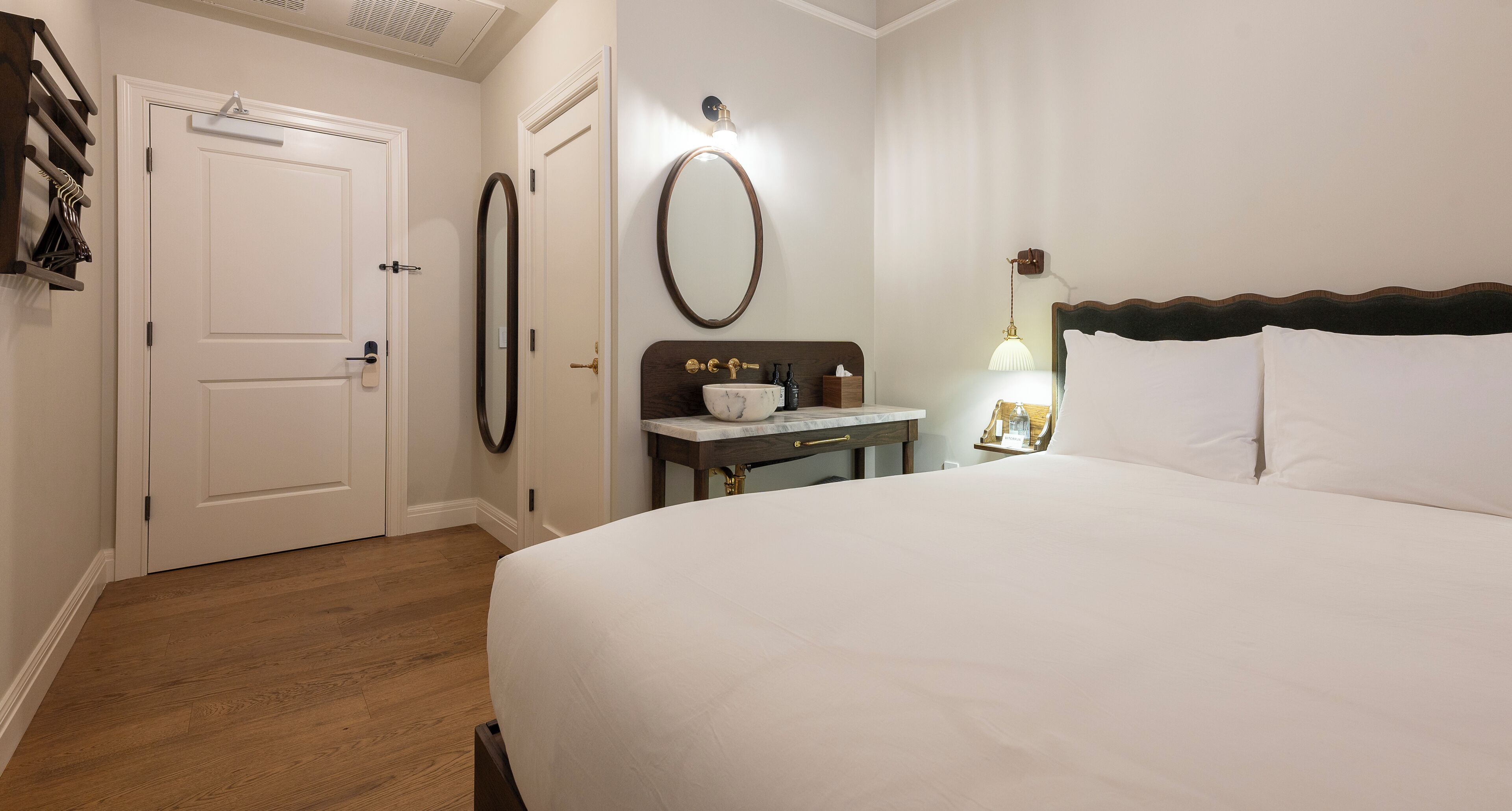 A hotel room with a bed, a nightstand, a mirror, a door, and a wooden floor.