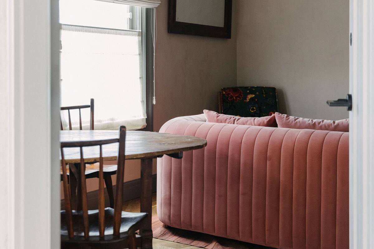 Et værelse med en lyserød sofa, et træspisebord med stole, et vindue med gardiner og et billede i ramme på væggen.