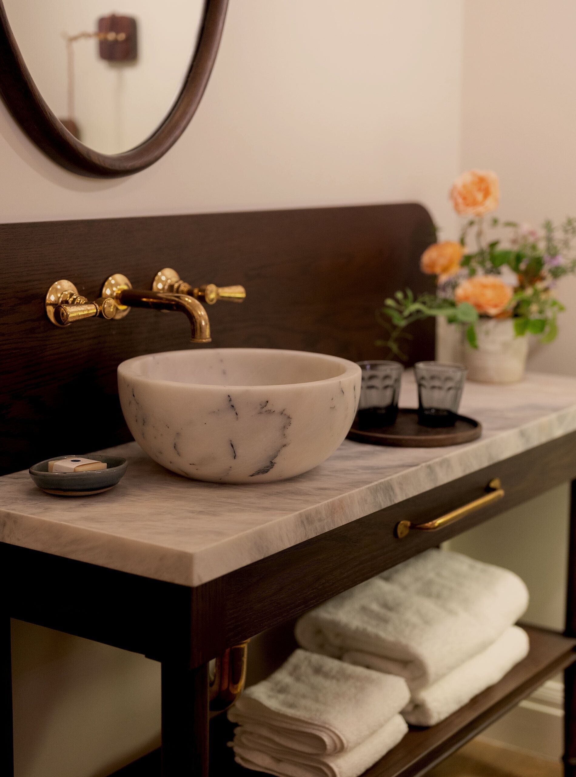 A bathroom with a marble sink, gold faucet, and a mirror above it. There is a vase with flowers on the counter and towels in a wooden cabinet below.