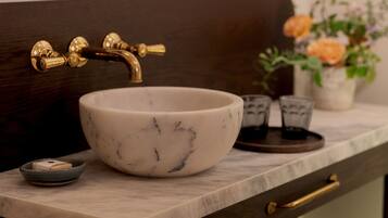 A bathroom with a marble sink, gold faucet, and a mirror above it. There is a vase with flowers on the counter and towels in a wooden cabinet below.