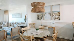 A cozy living room with a round glass table, wicker chairs, a wicker chandelier, and beach-themed artwork on the walls.