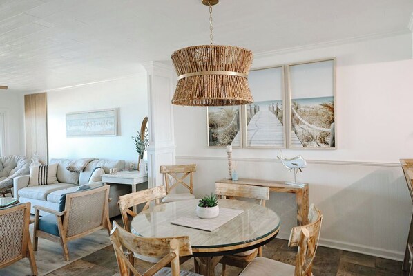 A cozy living room with a round glass table, wicker chairs, a wicker chandelier, and beach-themed artwork on the walls.
