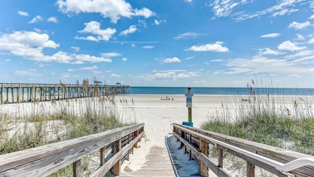 A wooden boardwalk leading to a beach with a pier in the background.