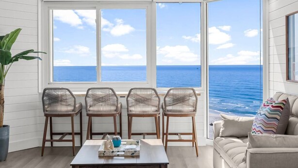 Una habitación con un amplio ventanal con vista al océano, sillas de mimbre, una mesa con una bandeja y un sofá con un cojín de colores.