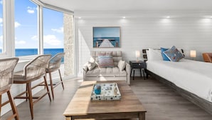 Habitación de hotel moderna con un amplio ventanal que ofrece vistas al océano, un sofá, una cama y una mesa de centro con una bandeja.