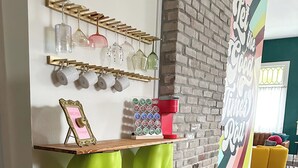 Una habitación con pared de ladrillo, un letrero colorido que dice 'Let the Good Times Roll', una cafetera y una estantería con vasos colgantes y un marco de fotos.