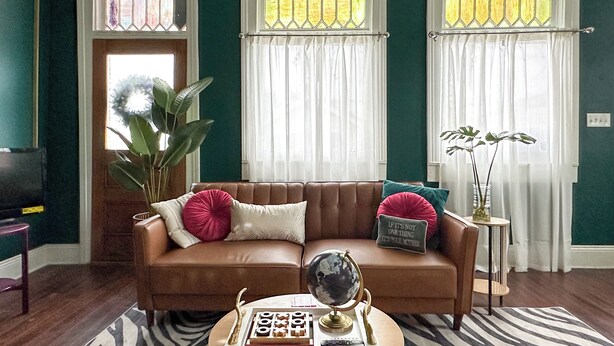 Una sala de estar con un sofá de cuero marrón, un globo terráqueo y una mesita con libros y un control remoto. Hay dos ventanas con cortinas, una planta y un televisor de pared.