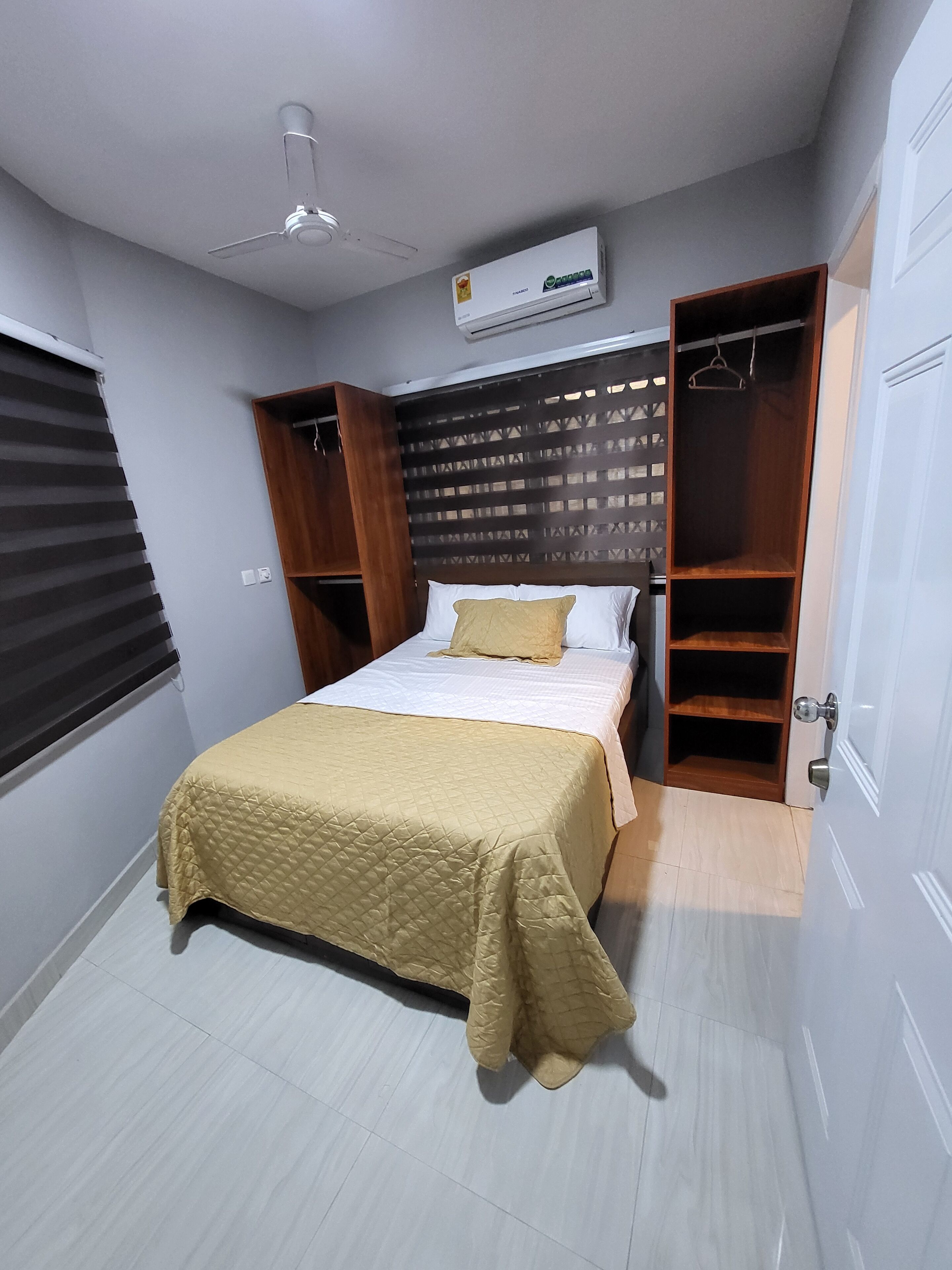 A bedroom with a bed, a ceiling fan, an air conditioning unit, a wooden wardrobe, and a window with blinds.