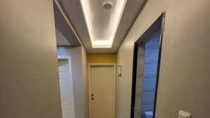 Un couloir étroit avec un luminaire au plafond, une porte au fond et un interrupteur fixé au mur.