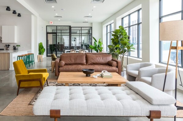 A modern living room with a brown sofa, yellow armchair, and white ottoman. There is a wooden coffee table with a bowl and books. The room has large windows, a plant, and a lamp.