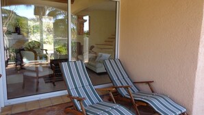 A sunlit patio with two striped lounge chairs and a sliding glass door leading to an interior space with a sofa and a dining table.